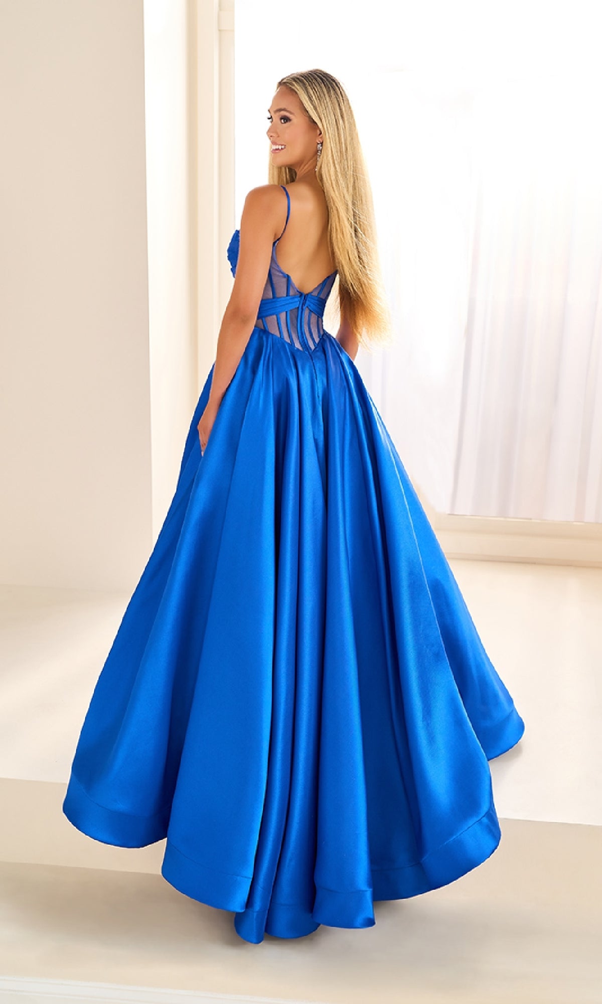 A woman wears the Sheer-Corset Prom Ball Gown: Ellie Wilde EW36073, a bright blue floor-length dress with a sweetheart neckline and strappy sheer back, as she turns and smiles in a sunlit room with white walls and large windows.